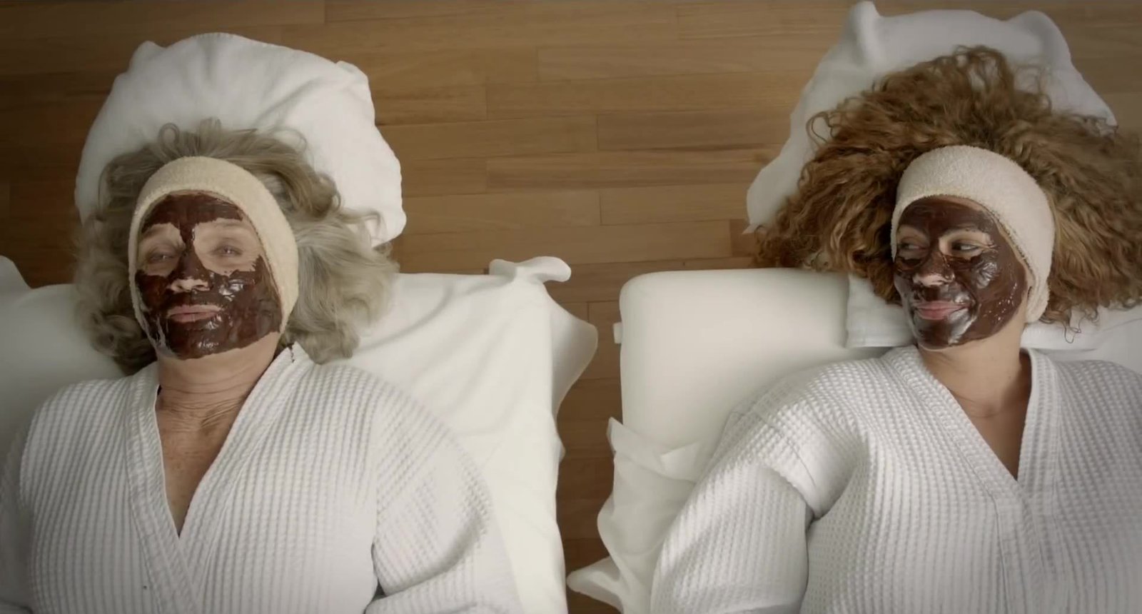 Two women with chocolate face masks lying in bed